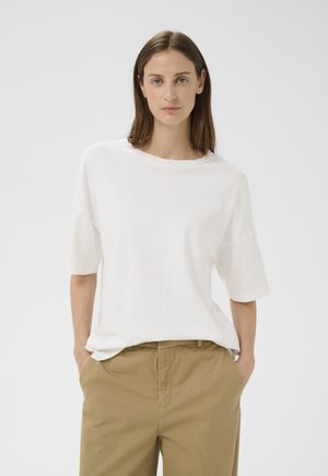 Donna con capelli castani lisci, che indossa una maglietta bianca a maniche corte e pantaloni kaki, in piedi con le mani nelle tasche contro uno sfondo semplice.