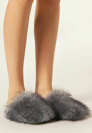 Pantofole grigie in peluche con una forma arrotondata, realizzate in materiale morbido, con dettagli soffici e pelosi attorno alle aperture.