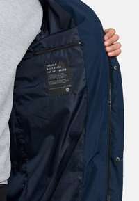 Marineblauwe jas met een gladde, glanzende binnenkant. Bevat een ritszak en een label met de tekst "Duurzame dagelijkse goederen voor elk seizoen."