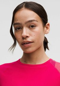 Ružová športová blúzka s okrúhlym výstrihom a priesvitnými sieťovinovými akcentmi na pleciach. Hladká textúra, priliehavý strih, ktorý zdôrazňuje tvár modelky.