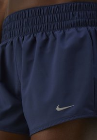 Primo piano di una persona di pelle scura che indossa pantaloni sportivi Nike blu navy con vita elasticizzata e logo Nike riflettente vicino all'orlo.
