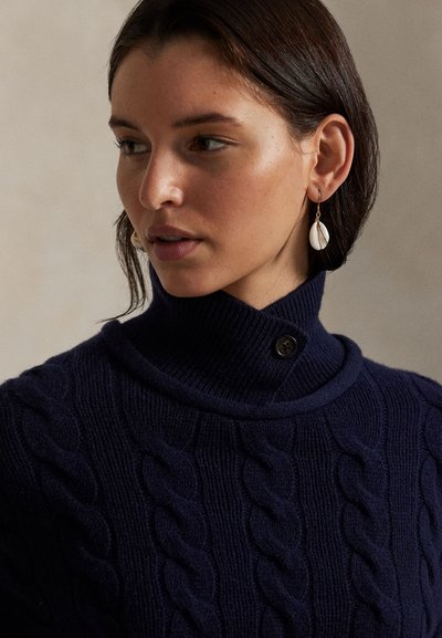 Νεαρή γυναίκα με σκούρα μαλλιά φοράει μπλε σκούνο πλεκτό πουλόβερ με ζιβάγκο και λευκά σκουλαρίκια με σχέδιο κοχυλιού, κοιτάζει ελαφρώς προς τα αριστερά.