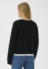 Fekete kötött pulóver kábelmintákkal, kerek nyakkal és bordás szegéllyel. Fehér felső fölött viselve, kék farmerral párosítva. Hátsó nézet látható.