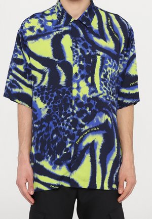 Lyhythihainen, napitukseton paita, jossa kirkkaan sininen, keltainen ja musta abstrakti eläinprintti, puettuna henkilölle, jolla on mustat housut.