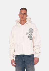 Lyse beige zip-up hoodie i blødt stof med en stor hætte, der har tre runde grafiske patches på venstre side og forlommer.