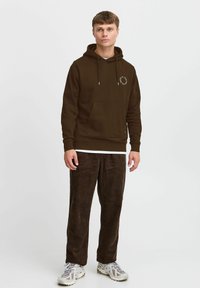 Brun hættebluse med kængurulomme og logo-detalje på brystet, parret med mørke kordfløjl bukser og grå sneakers.