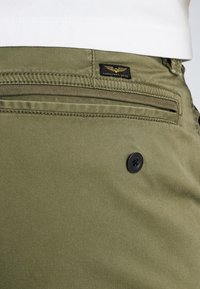 Olivgrüner Baumwollgemisch-Stoff mit flachem Frontteil, Seitentasche und schwarzem Knopf. Über der Tasche befindet sich ein schwarzes Etikett mit einem goldenen Logo.