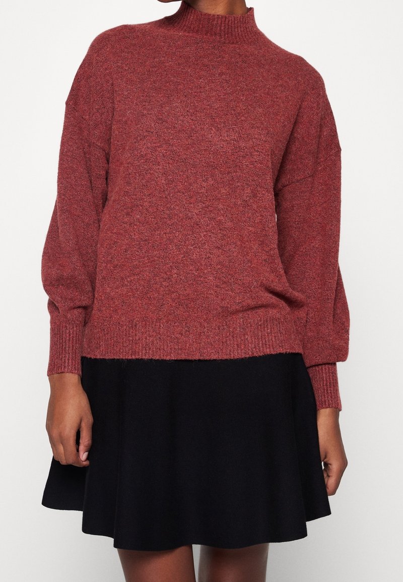 Donna che indossa un maglione lungo a maniche con trama di colore rosso scuro abbinato a una gonna nera semplice, in piedi contro uno sfondo bianco.