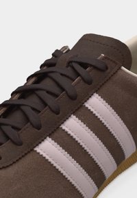 Καφέ σουέτ αθλητικό παπούτσι με σκούρα καφέ κορδόνια και τρεις ανοιχτό ροζ ρίγες στο πλάι, με άκρη σόλας από καουτσούκ.