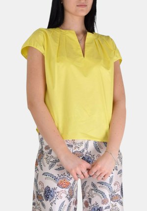 Blusa gialla a maniche corte con scollo a V e design plissettato. Abbinata a pantaloni chiari con motivo floreale, caratterizzati da accenti blu e arancioni.