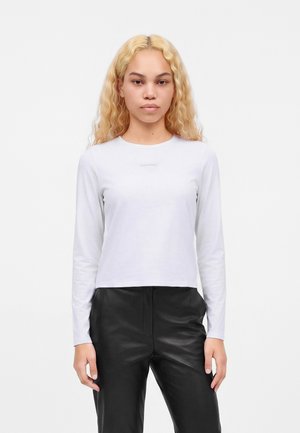 Femme aux longs cheveux blonds bouclés porte un haut blanc uni à manches longues et un pantalon en cuir noir sur fond blanc.