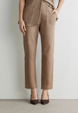 Kvinde iført brune bukser med lige ben og forlommer foran samt mørkebrune spidse hæle, stående op ad en lys væg.