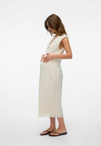 Robe de maternité en tricot texturé crème avec un décolleté en V et sans manches, portée avec des tongs noires, mettant en valeur une silhouette ajustée.