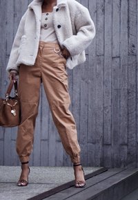 Veste en fausse fourrure beige, pantalon cargo tan avec poignets élastiques et sandales à talons. Un sac à main marron avec des détails en chaîne complète la tenue.