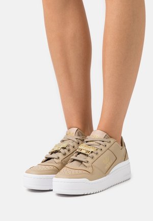 Baskets basses Adidas beiges avec semelles blanches et accents dorés portées sur des jambes nues contre un fond blanc.