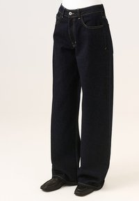 Szerokie ciemnoniebieskie dżinsy z zapięciem na guzik, kontrastowym przeszyciem i dwiema przednimi kieszeniami. Tekstura tkaniny wydaje się solidna i trwała.