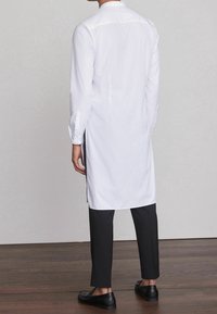 Camisa larga blanca con cuello, aberturas laterales y puños con botones, combinada con pantalones negros y zapatos negros sin cordones sobre un suelo de madera.