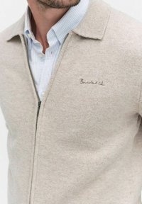 Beige zip-up cardigans i mjukt stickat material, med krage och subtil broderidetalj på bröstet. Bärs över en ljusblå skjorta.