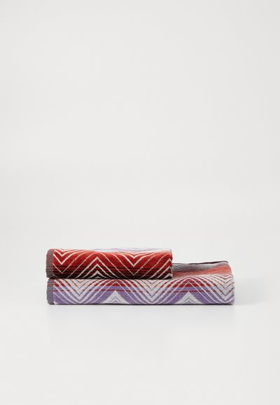Twee opgerolde handdoeken met zigzagpatronen. De bovenste handdoek is rood, wit en donkergrijs; de onderste handdoek is lichtpaars, wit en donkergrijs.