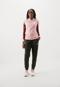 Rožinė zip-up striukė su tamsiai raudonomis rankovėmis, derinama su juodomis sportinėmis kelnėmis. Striukė turi logotipą, o sportbačiai yra rožinės spalvos. Lygi tekstūra.