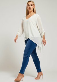 Vit, flödande blus med V-ringning och långa, uppvivna ärmar, matchad med smala blåjeans och nakna remsandaler med klack. Slät textur, asymmetrisk nederkant.