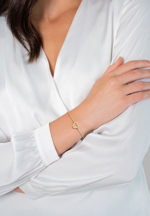 Pulsera de oro con un charm en forma de corazón, llevada en una muñeca. La cadena es delicada y los eslabones son pequeños, sobre una blusa blanca.