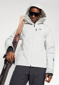 Casaco de ski branco à prova d'água com capuz, frente com fecho e detalhes pretos. O modelo segura esquis, usando óculos de sol escuros e luvas pretas.