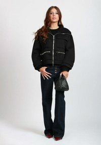 Giacca corta nera con cerniere argento, due tasche frontali, abbinata a jeans scuri a zampa d'elefante e una piccola borsa nera.