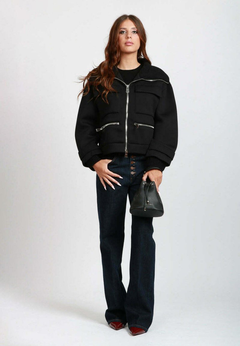 Giacca corta nera con cerniere argento, due tasche frontali, abbinata a jeans scuri a zampa d'elefante e una piccola borsa nera.