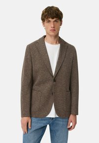 Brun tekstureret blazer med to forlommer, enkelt knaplukning og nakkerevers, båret over en hvid skjorte og blå jeans.