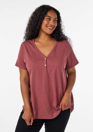 Frau mit lockigem Haar, die ein kurzärmliges rosafarbenes, geknöpftes V-Ausschnitt-Shirt trägt, lächelt und schaut nach links.