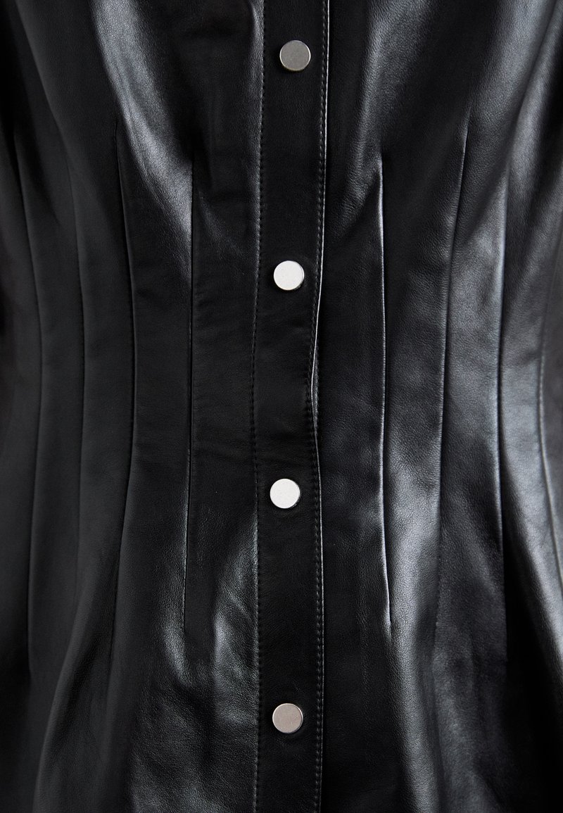 Corset en cuir noir avec des plis verticaux, featuring des boutons-pression en argent sur le devant et une texture lisse et brillante.