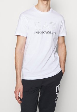 Mann trägt ein weißes Emporio Armani EA7 T-Shirt und schwarze Hose mit EA7-Logo am Oberschenkel vor einfarbigem Hintergrund.