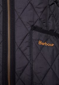 Czarna pikowana kurtka z brązowym zamkiem i haftowanym logo "Barbour" w kolorze pomarańczowym. Posiada boczną kieszeń z czarnym wykończeniem.