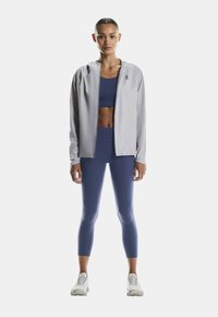 Hellgraue, zipfarbene Sportjacke über einem dunkelblauen Cropped-Top und passenden Leggings, mit einer figurbetonten Passform und dehnbarem Material. Weiße Sneaker runden den Look ab.