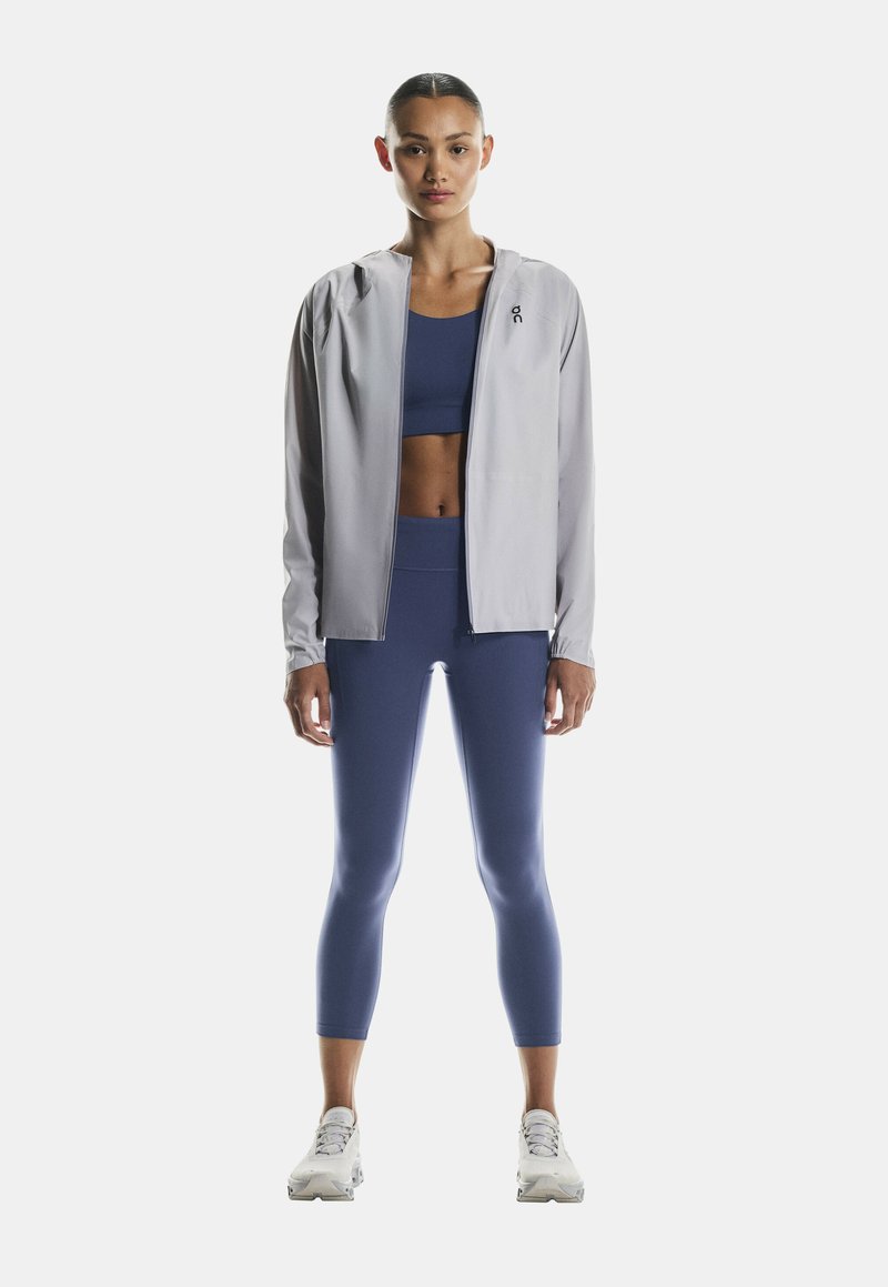 Hellgraue, zipfarbene Sportjacke über einem dunkelblauen Cropped-Top und passenden Leggings, mit einer figurbetonten Passform und dehnbarem Material. Weiße Sneaker runden den Look ab.