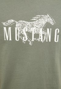 Illustration blanche usée d'un cheval au galop au-dessus du mot « MUSTANG » en grandes lettres capitales sur un tissu vert olive.