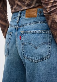 Άτομο που φοράει μπλε τζιν παντελόνι Levi's και καφέ καρό πουκάμισο, εμφανίζοντας τις πίσω τσέπες και την δερμάτινη ετικέτα της μάρκας Levi's.