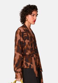 Bruine en zwarte blouse met patroon, lange mouwen en een gedrapeerde voorkant, gemaakt van een soepele stof. Geaccentueerd met een gele handtas.