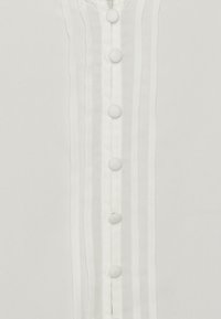 Detailní záběr na bílý oděv s vertikálním řádkem malých bílých knoflíků potažených látkou a jemným vertikálním prošíváním.