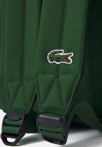 Sac à dos vert en tissu textur é, avec des bretelles ajustables et un clip noir. Comprend un logo de crocodile brodé distinctif.