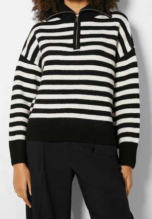 Sweter dzianinowy w czarno-białe paski z kołnierzem na półzamek, noszony z czarnymi spodniami, widok od szyi do bioder.