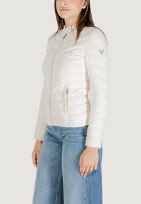 Giacca bianca imbottita con colletto alto, chiusura con zip e tasche laterali. Caratterizzata da una texture morbida con un leggero lucido, vestibilità avvitata e dettaglio del logo.