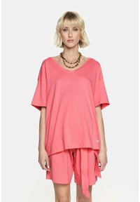 Różowa bawełniana bluzka z dekoltem w serek i krótkimi rękawami, zestawiona z dopasowanymi szortami z bokami wiązanymi węzłem. Gładka tekstura i luźny krój.
