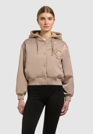 Young woman wearing a beige buttoned hoodie with embroidered logo on chest and black leggings, standing against a plain background.