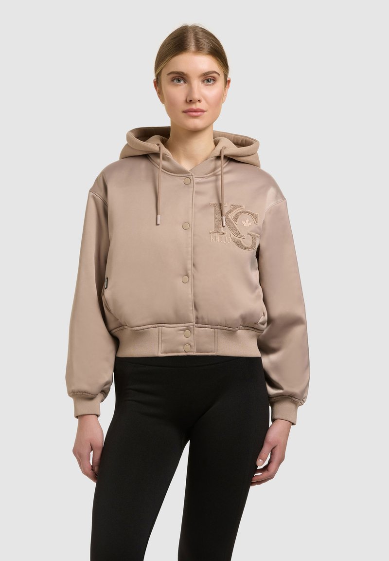 Jeune femme portant un sweat à capuche beige boutonné avec un logo brodé sur la poitrine et des leggings noirs, debout devant un fond uni.