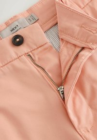 Pantalon rose clair avec une fermeture éclair ouverte, un bouton marron, et une étiquette de marque visible indiquant la taille 34R et le texte "Coupe droite".