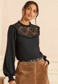 Fekete hosszú ujjú blúz csipkedíszítéssel a galléron, fodros szegélyekkel és sima textúrával, barna velúr szoknyával és fekete övvel párosítva.