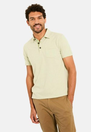 Hellgrünes Polohemd mit einer kleinen Brusttasche, zwei dunklen Knöpfen am Kragen und kurzen Ärmeln. Kombiniert mit beigen Hosen.