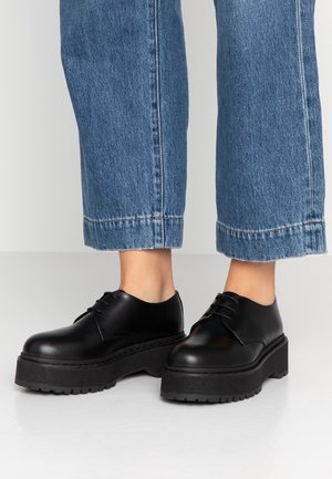 Schwarze Leder-Plateauschuhe mit Schnürung, getragen zu blauen, gekürzten Jeans auf weißem Boden und Hintergrund.